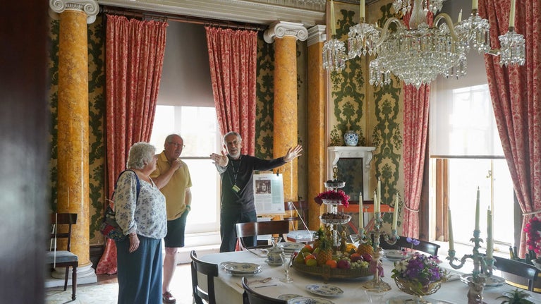 A volunteer talking to two visitors stood in front of a big window and looking down a dinning table